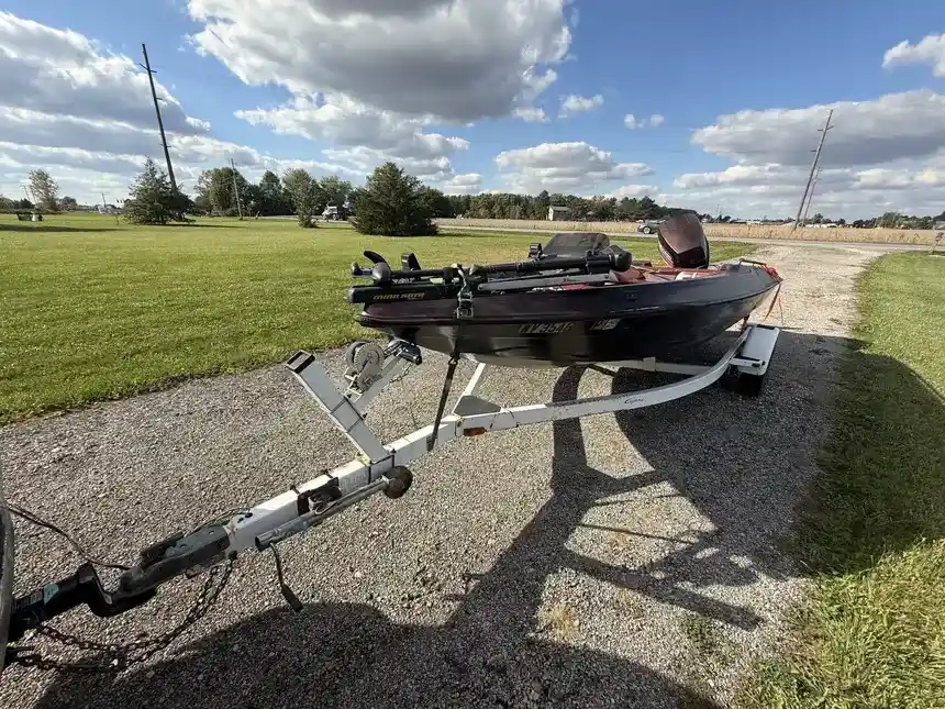 Black fishing boat with trolling motor on a single-axle trailer, parked on gravel next to a grassy field.