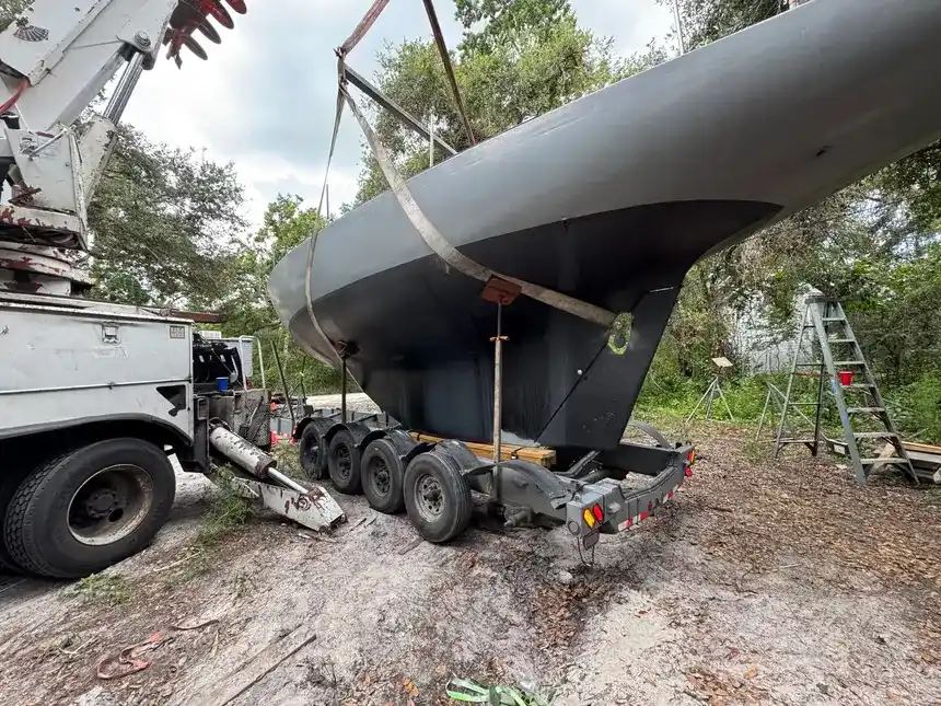 Heavy-duty crane lifting a large, dark gray boat hull onto a multi-axle trailer in a wooded yard.